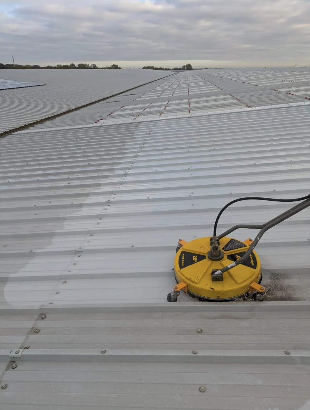 Rotary surface cleaner on a large warehouse roof, with a clean stripe showing against the dirty sheets